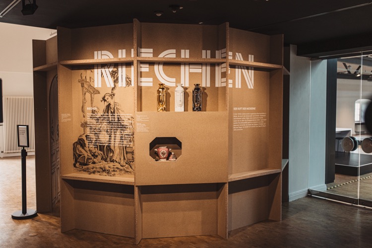 Kabinett "Riechen" in der der Sonderausstellung Sense&Sensibility im Museum Schloss Fürstenberg. Foto: Claudia Warneke Blick auf ein vieleckiges Kabinett aus rohen Pressholzplatte. An der Wand des Kabinetts steht in großen, weißen Buchstaben "Riechen". In eine Wand ist ein Regal eingelassen, auf dem moderne und historische Duftvasen stehen.